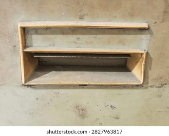 A rectangular, single slot yellow plastic letterbox or pass through set horizontally into a heavily stained and aged concrete wall. The exterior shows significant wear, dirt, and corrosion, with rust stains streaking down the rough concrete surface from the edges of the box. A small, rusty metal flap is partially visible at the back of the slot opening. The overall aesthetic is one of urban decay, age, and neglected maintenance. The photograph is taken head on with natural lighting, focusing on the textures of the old plastic and weathered concrete.