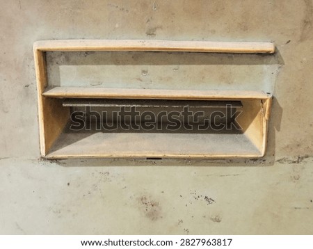 A rectangular, single slot yellow plastic letterbox or pass through set horizontally into a heavily stained and aged concrete wall. The exterior shows significant wear, dirt, and corrosion, with rust stains streaking down the rough concrete surface from the edges of the box. A small, rusty metal flap is partially visible at the back of the slot opening. The overall aesthetic is one of urban decay, age, and neglected maintenance. The photograph is taken head on with natural lighting, focusing on the textures of the old plastic and weathered concrete.