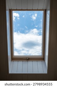 A rectangular skylight window set into a white paneled ceiling, revealing a bright blue sky with scattered white clouds through slightly dirty glass.