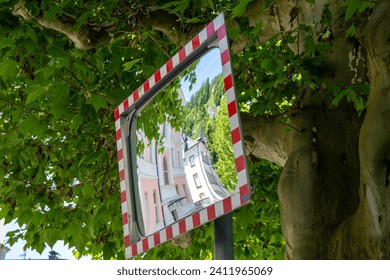 A rectangular traffic mirror with a red and white frame and a distorted reflection of a city. The mirror is mounted in front of a tree with green leaves and the sun is shining.