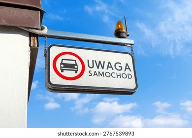 A rectangular warning sign hangs from a metal beam against a bright blue sky, featuring a red-circled car icon and the Polish text 