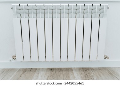 A rectangular white radiator sits on a hardwood floor next to a white wall. The flooring has a wood stain finish, creating a cozy and elegant look