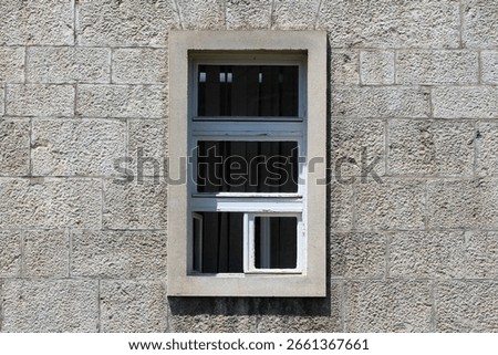 A rectangular, white-framed window with multiple divided glass panes and a slightly open lower panel is set in a textured stone building facade