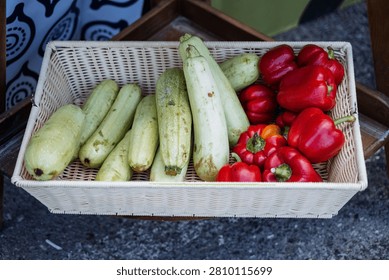 Rectangular wicker basket filled with a row of pale green zucchini and a cluster of glossy red bell peppers basket sits on rough concrete pavement next to a patterned fabric