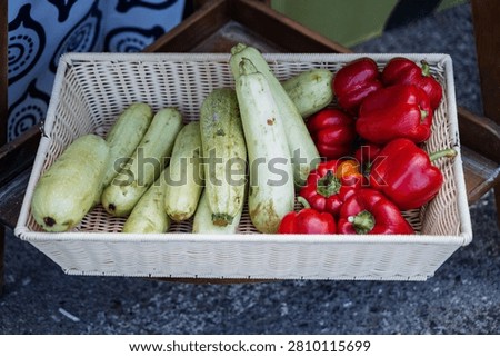 Rectangular wicker basket filled with a row of pale green zucchini and a cluster of glossy red bell peppers basket sits on rough concrete pavement next to a patterned fabric