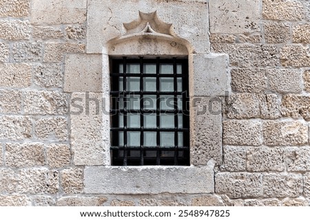 Rectangular window with black colored metal grille on an old beige colored stone wall. From the Peace window series.