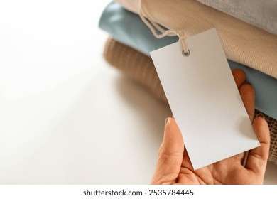 Recyclable product concept. A stack of neutral toned knitted clothes made of natural materials lies on the table. A hand holds a blank white label. 