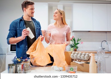 Recycling. Responsible young couple putting empty plastic and paper in garbage bags while looking at each other near the table filled with waste - Powered by Shutterstock