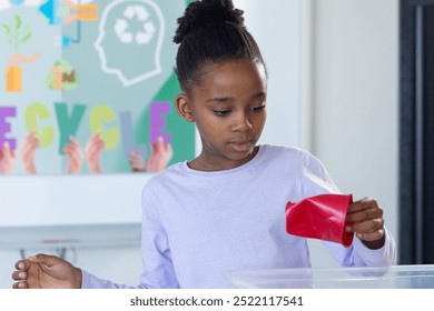 Recycling in school, african american girl placing red paper into recycling bin, focusing on task. environment, sustainability, education, eco-friendly, waste management, responsibility - Powered by Shutterstock