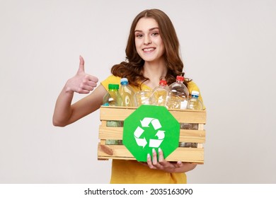 Recycling, waste sorting and sustainability concept. Smiling young female in yellow t-shirt holding box with plastic bottles and showing thumb up. Indoor studio shot isolated on gray background. - Powered by Shutterstock