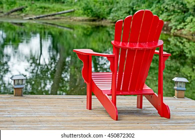 Red Adirondack Chair At A Lake - Also Known As A Muskoka Chair In Canada, After The Ontario Lake Of The Same Name.  These Chairs Are An Iconic Symbol For Successful Retirement To A Woodland Cottage.