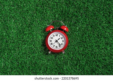 Red Alarm Clock On Green Turf Grass Background. Half Past One O'clock. Copy Space, Game Time Management Concept. Opening Or Closing Hours. Ecological Sustainable Business Banner