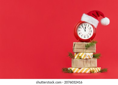 Red alarm clock in Santa Claus hat on wrapped gift boxes decorated fir branches and gold cones. Red background. Concept of coming Christmas and New Year, holiday sales. Space for text.