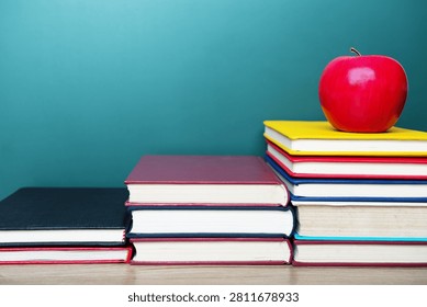 A red apple is placed on the top of a pile of books, with a blackboard in the background.