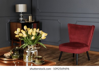 Red Armchair In Dark Grey Living Room Interior With Lamp And Yellow Flowers On Table In Blurred Foreground