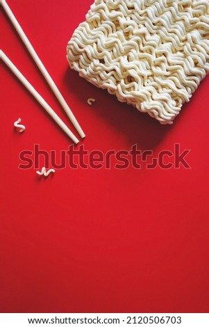 Red background with uncooked instant noodles or dried noodle block in typical block form and wooden chopsticks. Eating behavior, effects of instant noodles on human health. (top view, space for text)