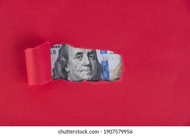 A Red Background, From Under Which A Portrait Depicted On A Hundred Dollar Bill Peeps.