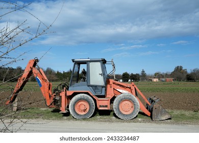Red backhoe with large wheels parked on edge of dusty road, surrounded by grass and trees on plowed land background