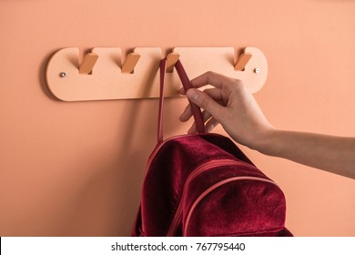 Red Backpack Is Hanging On The Metallic Orange Hanger On The Same Color Wall Indoors. Hand Is Holding The Backpack's Strap. Closeup Horizontal Photo. 