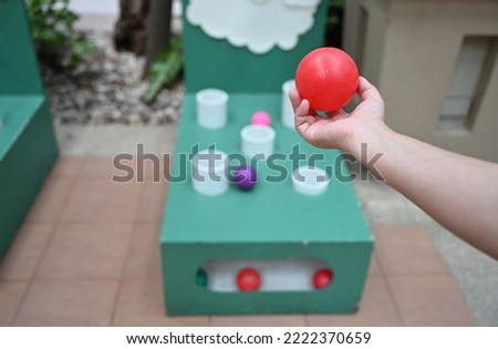 Red ball in right hand in preparation for throwing into the white pipe(hole) with some of the ball falling into the wood box. Throwing the ball into the gap is carnival tossing game. practice accuracy