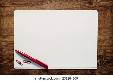 Red Ballpoint Pen And Paperclips Lying On A Stack Of Blank Sheets Or Pages Of White Paper With Copyspace For Your Text On A Wooden Desk, View From Above