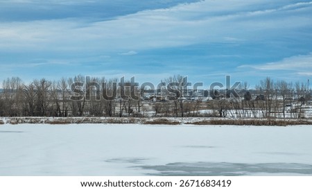 A red barn between trees on a cloudy winter day in Colorado with frozen lake in foreground