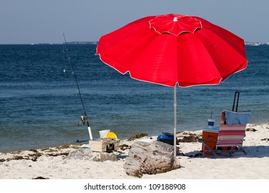 Red Beach Umbrella Shades A Spot In The Sand With A Beach Chair, Cooler And Fishing Tackle.