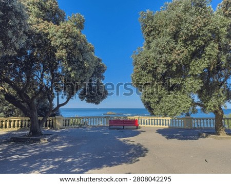 Red bench overlooking the Atlantic Ocean at Praia da Luz in Foz Porto Portugal framed by large trees and a stone balustrade under clear blue skies