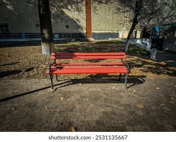 A red bench sits in front of a building with a tree behind it. The bench is empty and the leaves on the tree are falling - Powered by Shutterstock