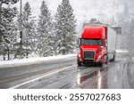 Red big rig commercial semi truck transporting cargo in dry van semi trailer running on the wet turning road with winter forest at snowing weather during a snow storm near Shasta Lake in California