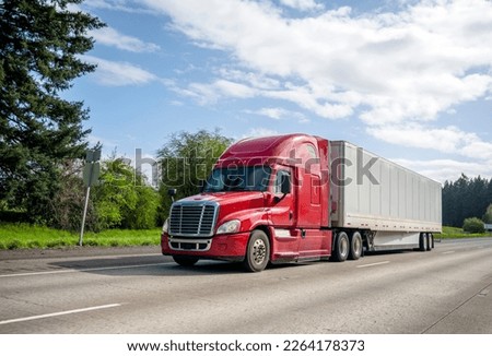 Red big rig popular professional reliable bonnet long haul semi truck transporting commercial cargo in dry van semi trailer moving on the straight wide highway with green trees on the background