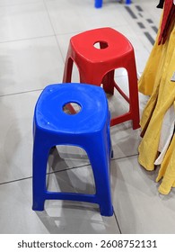 A red and blue plastic stool with circular seat holes stand side by side on a tiled floor, forming a playful contrast among colorful fabrics in a retail space.