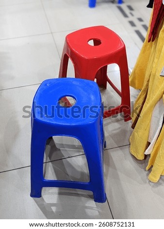 A red and blue plastic stool with circular seat holes stand side by side on a tiled floor, forming a playful contrast among colorful fabrics in a retail space.