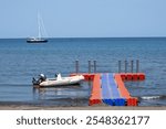 A red and blue pontoon pier on the beach a boat and yacht in the background. A floating plastic dock on a beautiful bay.