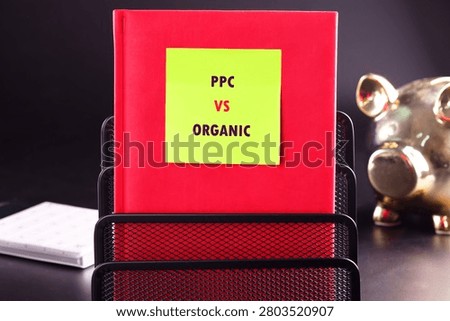 A red book titled PPC vs Organic is placed on a black holder. There is a gold piggy bank in the background. The setting is suitable for a marketing discussion.
