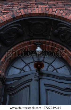 Red brick arch frames dark carved wooden double doors in Katowice, Poland. Semicircular fanlight, black stone reliefs, globe light, and red loudspeaker are present.