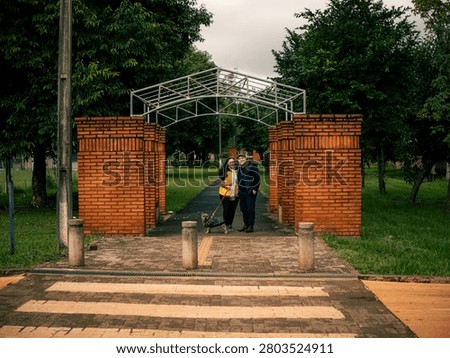 Red brick archway frames a metal gateway structure. Lush green trees fill the park background. Two people stand close under the arch. They smile, suggesting a warm, happy moment