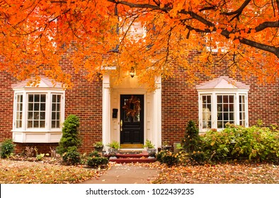 Red Brick House Entrance With Seasonal Wreath On Door And Porch And Bay Windows On Autumn Day With Leaves On The Ground And Hydrageas Still In Bloom - Colorful Foliage