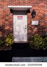 Red Brick Wall With White Door With A Security Sign. Security Camera Sign On An Entry Door Of A Commercial Business Building.