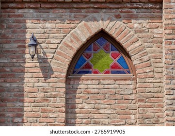 The red brickwork frames the window, creating a rustic contrast to the vivid blue and green hues of the glass. A classic lantern-style light fixture is mounted to the left