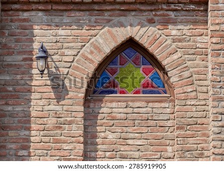 The red brickwork frames the window, creating a rustic contrast to the vivid blue and green hues of the glass. A classic lantern-style light fixture is mounted to the left