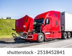 Red broken big rig semi truck with open hood and dry van semi trailer standing out of service on the road shoulder waiting for the towing truck or mobile road assistant for engine repair help