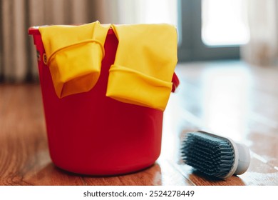 Red bucket with yellow gloves on hardwood floor, brush and mop in front - Powered by Shutterstock