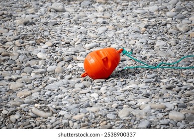 A red buoy is laying on the ground in front of a rocky shore. The buoy is tied to a rope, which is attached to a boat. The scene is calm and peaceful, with the buoy floating on the water - Powered by Shutterstock