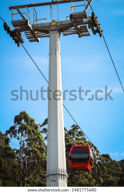 Red cab suspended by steel cable to carry\
people. aerial funicular, cable car. Cables support post, blue sky\
and trees in the\
background.