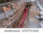 Red cables lie inside a deep earthen trench at a work site. Stacks of bricks and stone tiles border the excavation area along the road