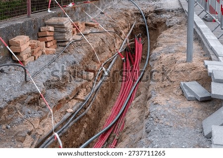 Red cables lie inside a deep earthen trench at a work site. Stacks of bricks and stone tiles border the excavation area along the road
