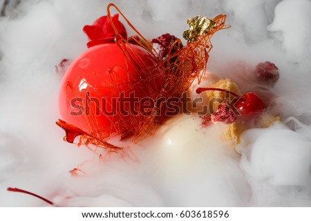 Red cake, yogurt spheres, raspberries, a caramel decoration and a fresh flower in steaming dry ice, close-up. Contemporary dessert.