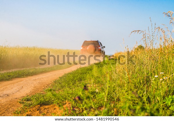 A red car is driving along a dirt road picking\
up a cloud of dust. Country road passing along the field. Clear\
sunny weather.