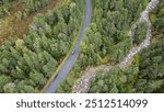 Red car driving on a winding road through a pine forest. Small creek with pebbles. Douglas fir and Scots Pine forest in the Cairngorms National Park. Drone shot in the Scottish Highlands, Scotland. 